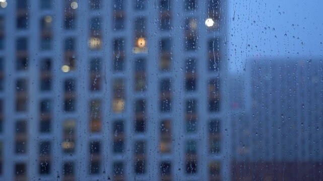 Rain drops flow down window glass on background of blurred windows house in big city on autumn evening. Rain in defocus window. Weather concept