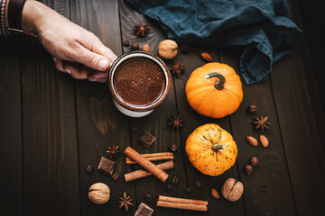 Male hand holding mug of hot chocolate, pumpkins and spices on rustic wooden background, cozy autumn scene