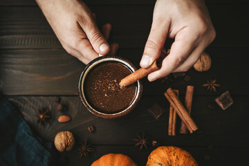 Top view of male hands holding mug of hot chocolate and cinnamon stick, pumpkins and spices on rustic wooden background, cozy autumn scene