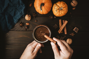 Top view of male hands holding mug of hot chocolate and cinnamon stick, pumpkins and spices on rustic wooden background, cozy autumn scene