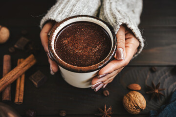 Female hands in sweater holding mug of hot chocolate drink on rustic wooden background, cozy seasonal scene