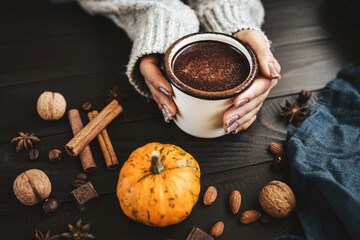 Female hands in sweater holding mug of hot chocolate, pumpkins and spices on rustic wooden background, cozy autumn scene
