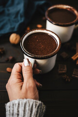 Female hand in sweater holding mug of hot chocolate, pumpkins and spices on rustic wooden background, cozy autumn scene