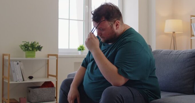 Tired fat overweight obese man holds forehead on couch. Male person with obesity shows headache, fatigue, stress, pain, migraine, exhaustion, and worry while sitting on sofa at home