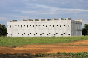 Foto sebuah bangunan beton berwarna putih dengan deretan jendela kecil yang terlihat sederhana dan minimalis. Bangunan ini berdiri di area padang rumput hijau dengan langit biru cerah di latar belakan