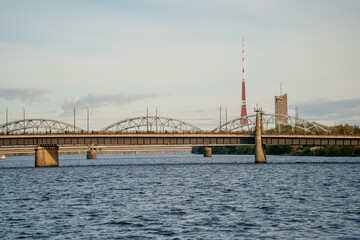 Fototapeta premium Riga skyline with steel railway bridge and television tower standing above the river, symbolizing connection between heritage, communication, and modern engineering in Baltic landscape harmony