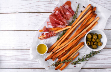 Grissini made of wheat flour with parmesan, rosemary and olive oil. Breadsticks served with olives and prosciutto on white background. Italian crispy snack. Top view