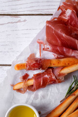Grissini made of wheat flour with parmesan, rosemary and olive oil. Breadsticks served with prosciutto on white background. Italian crispy snack