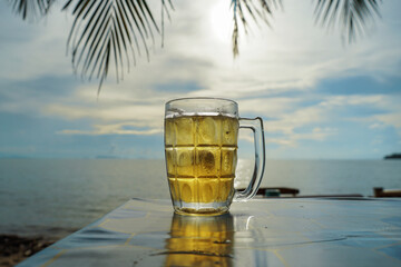 Icy cold beer mug with condensation rests on a wooden table. In the background, the sun sets over a peaceful beach. Alcohol beverage menu. Sunsets over the horizon by the ocean.