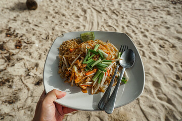 Top view of a delicious pad Thai dish served at a beachfront on beach sand in the background. A plate of food with pork and lime traditional Thai fried noodle dish.