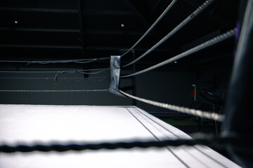 A boxing ring with white canvas and black ropes. The scene is dimly lit, emphasizing the structure...