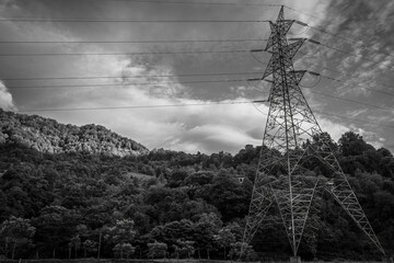 High-voltage electricity transmission tower standing in a lush green forested landscape with hills and dramatic clouds in the background