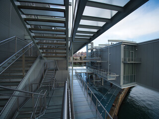 Modern Architecture Walkway Over Water - A metal and glass walkway with industrial design leads over water, reflecting the sky and clouds with a futuristic feel.