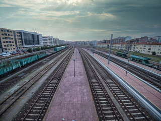 Fototapeta premium Busy Railroad Tracks in Urban Center - A high-angle shot of multiple railway tracks and platforms converging into the distance, with city buildings and cargo wagons under a dramatic, cloudy sky.