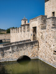 Obraz premium Modern Moat and Fortress in Spain - A fortified stone tower house stands surrounded by a square moat and a wide stone bridge, with a small fortified wall and entrance under a clear blue sky.