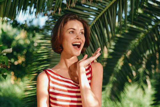 Fototapeta A cheerful woman in a striped sleeveless top stands outdoors among lush tropical plants, warmly lit by sunlight. She smiles brightly, radiating positive energy and relaxed confidence.