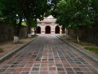 Naklejka premium Stone path leading to a royal temple in Hue - A low-angle shot of a traditional tiled pathway, flanked by trees and walls, leading to a multi-arched gate of a Vietnamese royal temple.