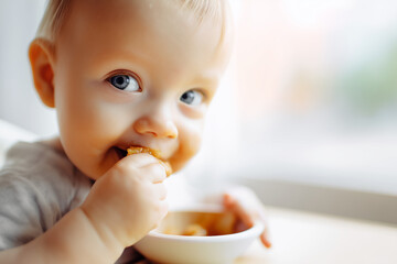 Close-up of a baby eating with hands from a bowl. Soft focus and natural lighting create a gentle, warm atmosphere. Copy space for text.
