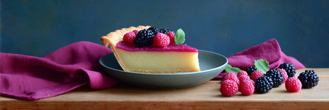 Slice of custard tart with berry topping served on a plate with fresh raspberries and blackberries on wooden table, close-up