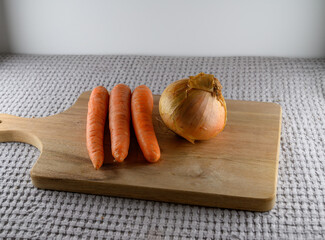 Freshly harvested carrots and a brown onion rest on a wooden cutting board. This scene captures the essence of meal preparation in a warm kitchen