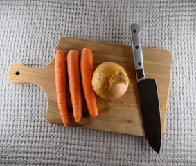 Fresh carrots and a large onion rest on a wooden cutting board alongside a sharp knife, ready for chopping and cooking delicious meals in a cozy kitchen setting