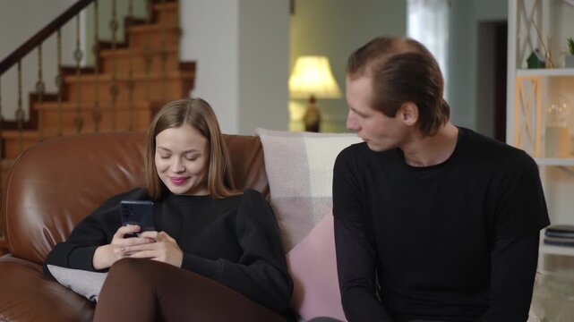 A young woman is focused on her smartphone with a smile, while her husband looks at her with concern. The scene shows distraction, neglect, and emotional distance in their family relationship at home.