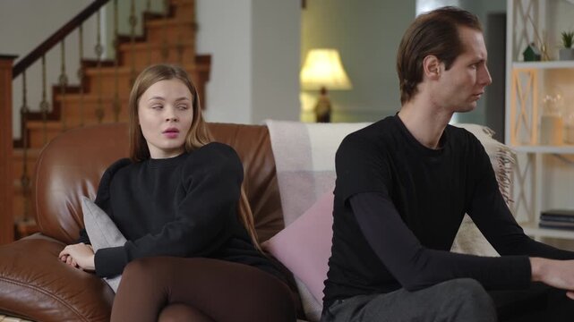 A man and woman sit on the sofa facing away from each other with serious expressions. The scene reflects silence, conflict, and emotional distance in their strained relationship at home.
