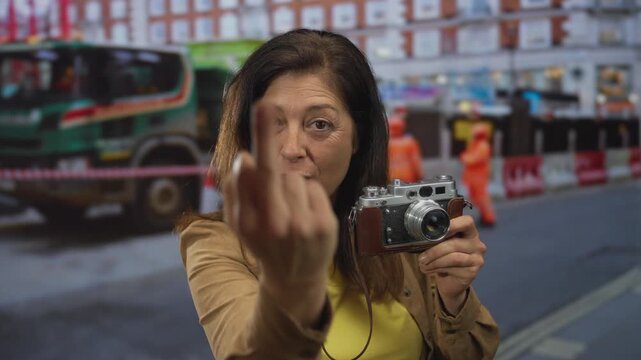 Woman holding vintage camera making middlefinger gesture in front of building; bold defiance attitude.