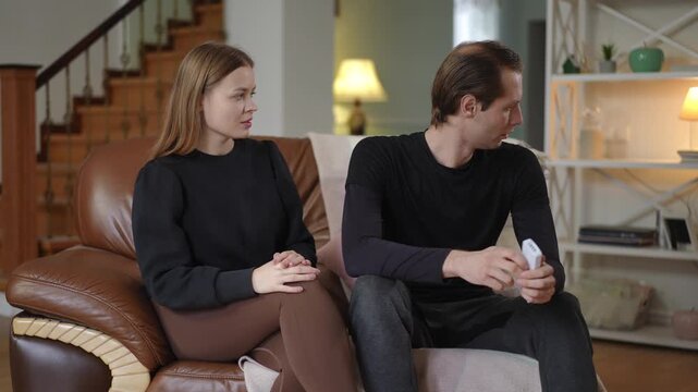 A woman looks at her husband with a conciliatory smile, trying to restore peace, while he sits with a serious expression, avoiding eye contact. The scene reflects tension, misunderstanding, and an