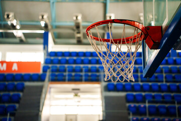Basketball hoop in empty indoor sports arena. Close-up of basketball hoop and net inside empty sports arena with blue seats in the background. © Evgenii
