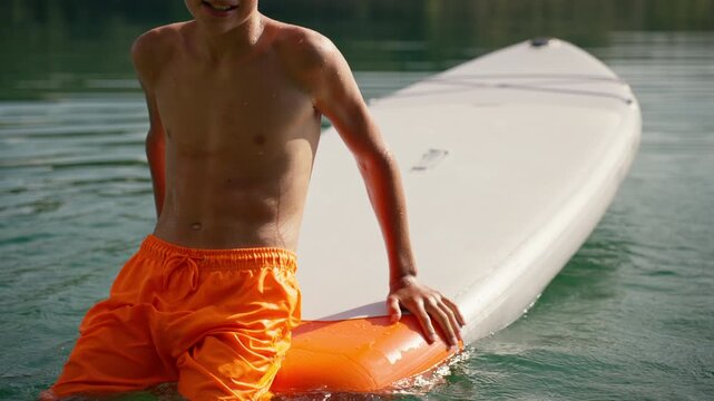 Energetic boy in orange shorts attempting to climb onto stand-up paddleboard on serene lake, practicing his
balance and enjoying sunny day during his summer vacation