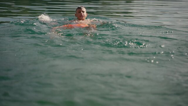 Cheerful young boy with short hair swimming in calm lake during sunny summer day, practicing backstroke and splashing water as he moves forward in refreshing green water