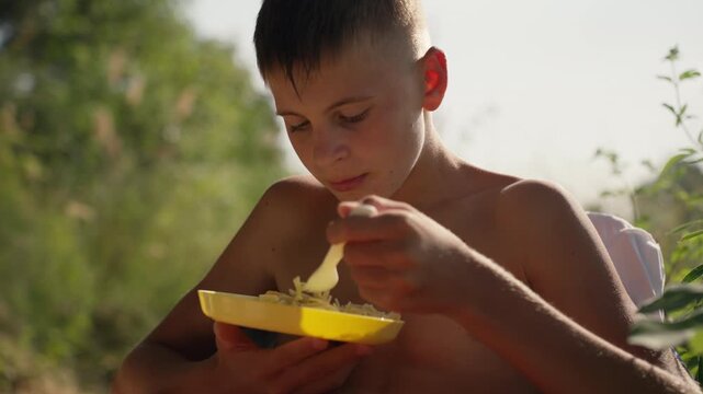 Young shirtless boy enjoy sunny outdoor picnic, eagerly savoring tasty meal on yellow plate while relaxing in warm summer park setting