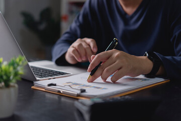 Businessman analyzing financial data report, hand with pen pointing at chart. Man working with laptop office desk, reviewing business performance, investment growth, and market strategy for success.