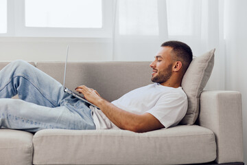 Relaxed man using a laptop while lounging on a comfortable couch, showcasing a modern lifestyle and cozy home environment filled with bright natural light.