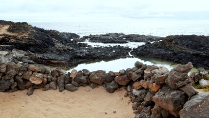 Volcanic rock pools providing a natural bathing area for naturist tourism, with the ocean in the background and a sandy beach foregrounded by a low stone wall