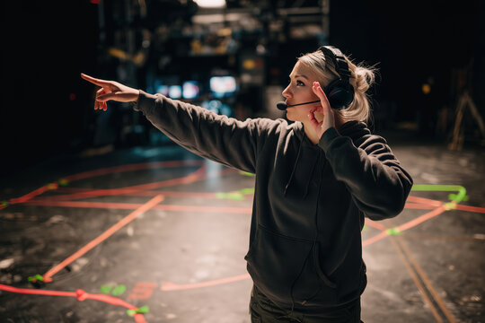 A blonde female production assistant, stage manager with headphones points on a dark stage. Woman directs during a show's filming with various technical markings on the floor. Live event production.