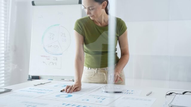 Businesswoman pointing at financial charts and graphs on a table, with a pie chart on a flip chart in the background. Business people concept