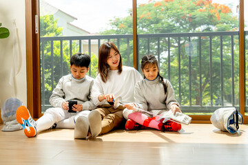 Asian mother, son and daughter as family sit on the floor in front of glass door with girl relax with smiling and boy enjoy the mobile phone.