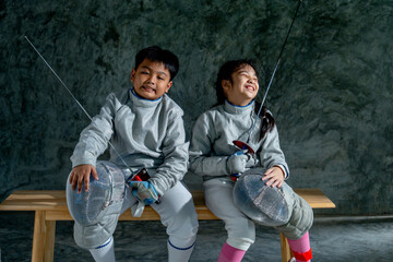 Asian brother and sister with fencing sport uniform sit beside on wooden bench and action of funny  also relax after practice for competition.
