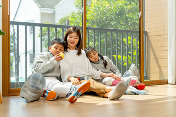Side view of Asian mother, boy and girl as family sit on the floor in front of glass door with girl sleeping and boy enjoy eat an apple after relax of practice in their house.