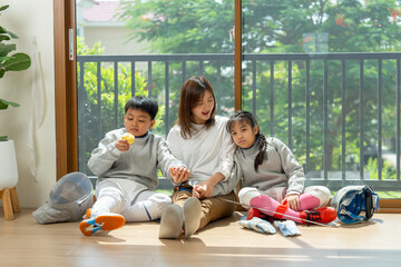 Front view of Asian mother, boy and girl as family sit on the floor in front of glass door with girl lean against mother and boy enjoy eat an apple after relax of practice in their house.