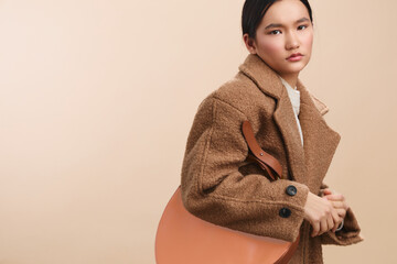 asian woman wearing a warm brown coat holds a large bag, portrait style with a confident pose. The neutral studio backdrop emphasizes chic street fashion and a modern emotional appeal.