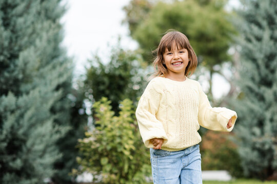 Smiling child girl 4-5 year old wearing knitted sweater and denim pants having fun in park over nature background outdoors. Looking at camera. Autumn season.