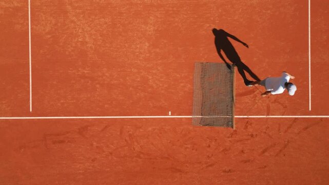 A bird's-eye view of tennis groundsman on a clay court, captured mid-action with a dynamic shadow. The setting emphasizes sports training and skill development, highlighting the textured clay surface 