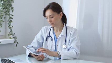 Confident female doctor wearing lab coat and stethoscope using digital tablet while sitting at desk in medical office, browsing patient information or conducting online consultation. Medicine concept