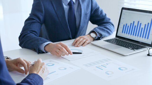 Business colleagues discussing financial data on a laptop and printed documents while analyzing charts and market trends. Business people and finance concept