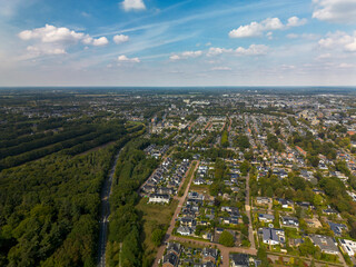 Aerial view of suburban neighborhood beside forest, with modern houses, organized streets, green spaces, and distant urban zone showcasing planned development and natural contrast.