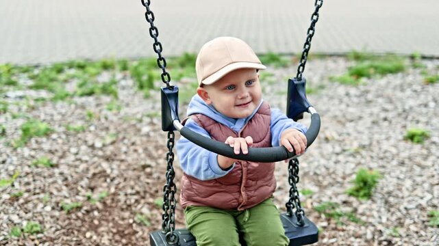 Happy toddler swings on a swing outside. A boy of 2 years old in a cap and a warm vest swings on a playground. The child has fun on a swing during a walk.