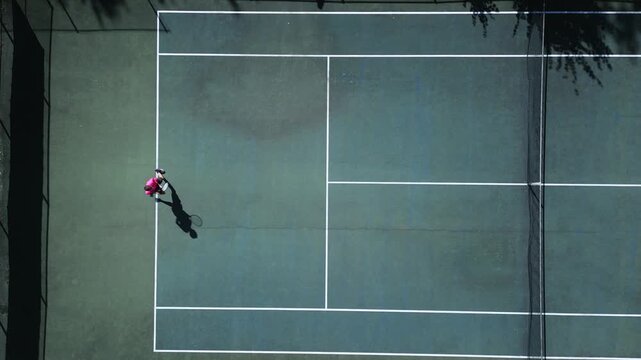 Tennis player in pink shirt practices on hardcourt, showcasing focused training and athleticism. Aerial shot captures dynamic motion and dedication in a professional sports setting.
